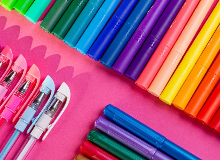 Hidrocores e canetas Tris dispostos sobre uma superfície rosa. Os itens estão organizados em fileiras, variando em cores.