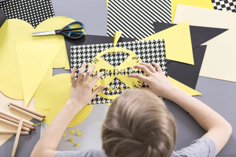 Criança criando colagem com papel colorido, tesoura e lápis em uma mesa.