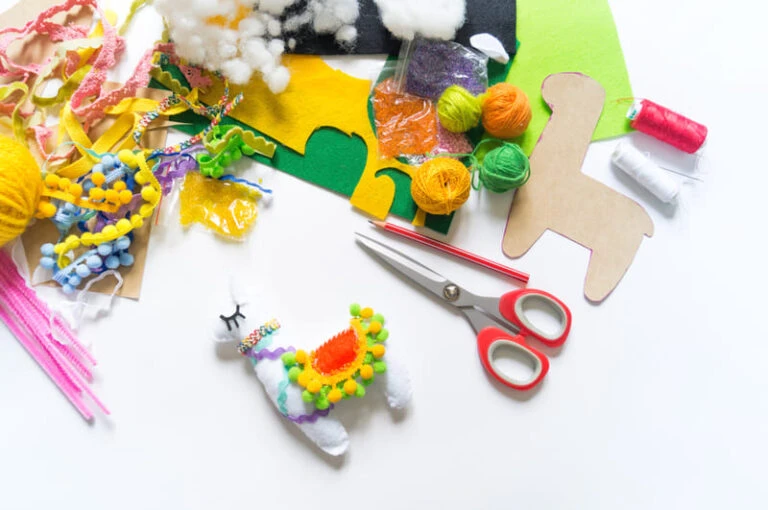Diversos materiais de artesanato, incluindo lã colorida, feltro, tesoura e um boneco de alpaca decorado, sobre uma mesa.