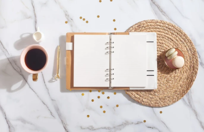 Imagem de um caderno aberto em uma mesa elegante ao lado de uma xícara de café e doces.