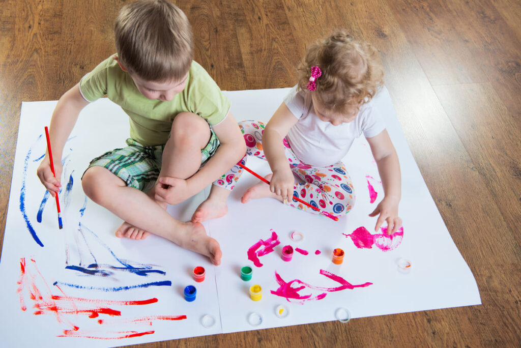 Duas crianças sentadas em cartolina pintando com tintas e pincéis.