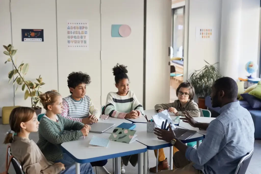 Grupo de crianças em uma atividade de aprendizado em sala de aula com um instrutor, rodeados de materiais escolares e tecnologia.