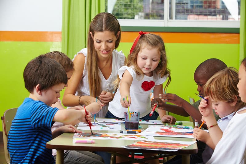 Grupo de crianças sentadas juntas criando pinturas e professora agachada ajudando