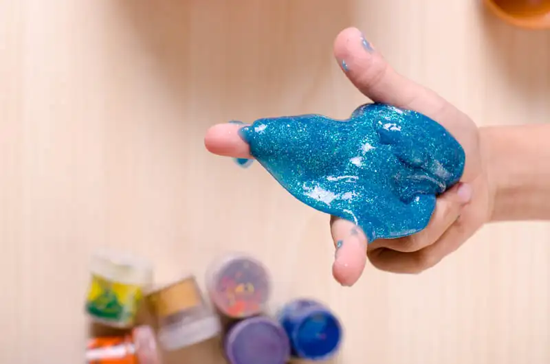 Mão de criança mostrando slime azul brilhante e potes de glitter ao fundo