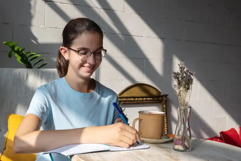 Jovem de óculos sorrindo, escrevendo em caderno na mesa com caneca e vaso de flores