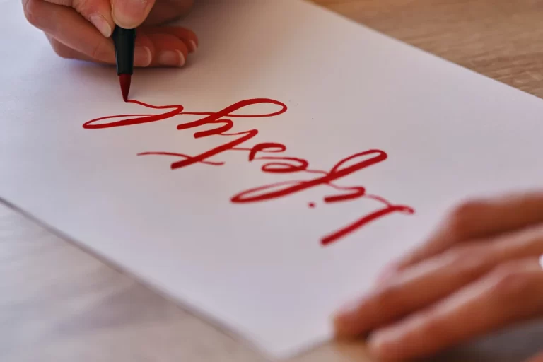 Uma mão segura uma caneta brush vermelha escrevendo em lettering em papel branco.