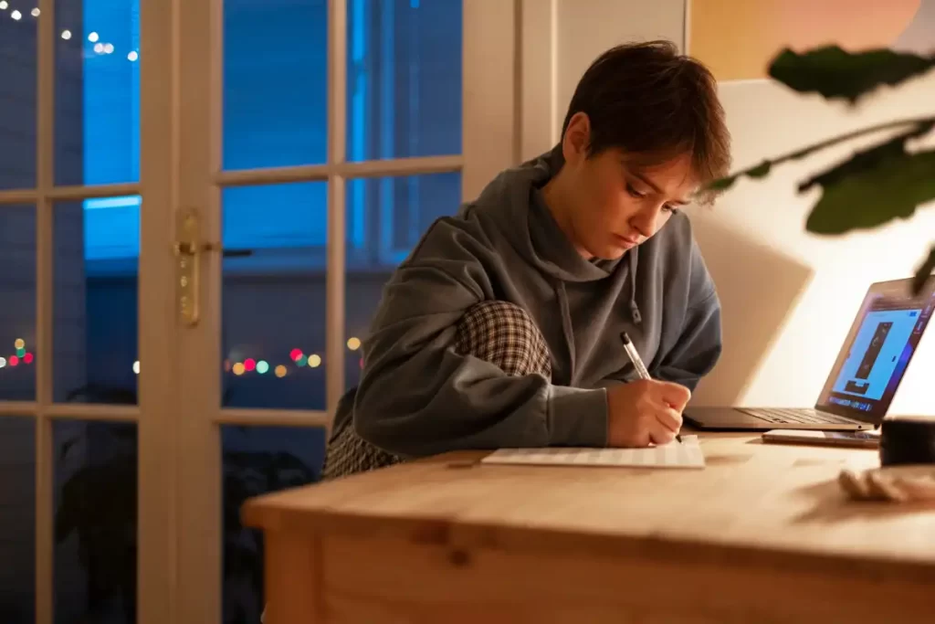 Jovem estudando em casa à noite, concentrado em suas anotações com um laptop ao lado.