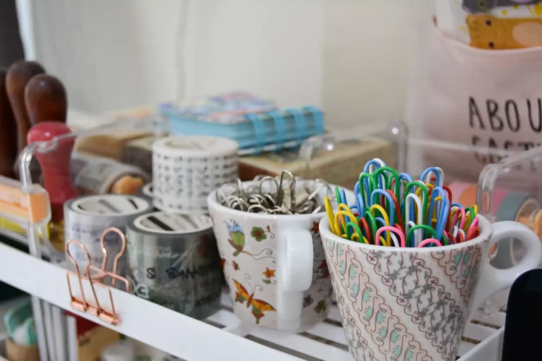 Imagem de organização de materiais de escritório com canecas, grampos coloridos e fitas adesivas em uma prateleira.
