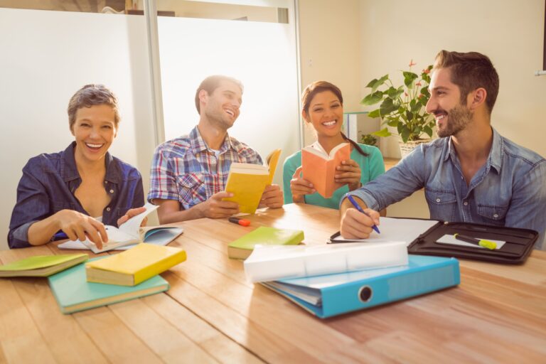 Grupo de pessoas estudando e discutindo em uma sala de aula com livros e material para faculdade ao redor, demonstrando colaboração e aprendizado.