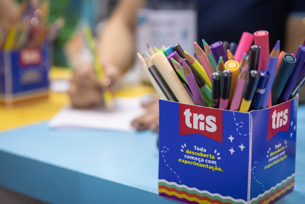 Conjunto de lápis coloridos e itens de papelaria Tris em uma caixa azul mensagem sobre experimentação, em ambiente escolar ou de entretenimento infantil.