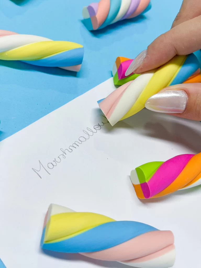 Mãos escrevendo na folha com borrachas coloridas em forma de doces, em um ambiente de escritório ou de estudos, com foco na atividade de apagar. Ideal para conteúdo sobre produtos de papelaria, organização escolar ou atividades criativas.