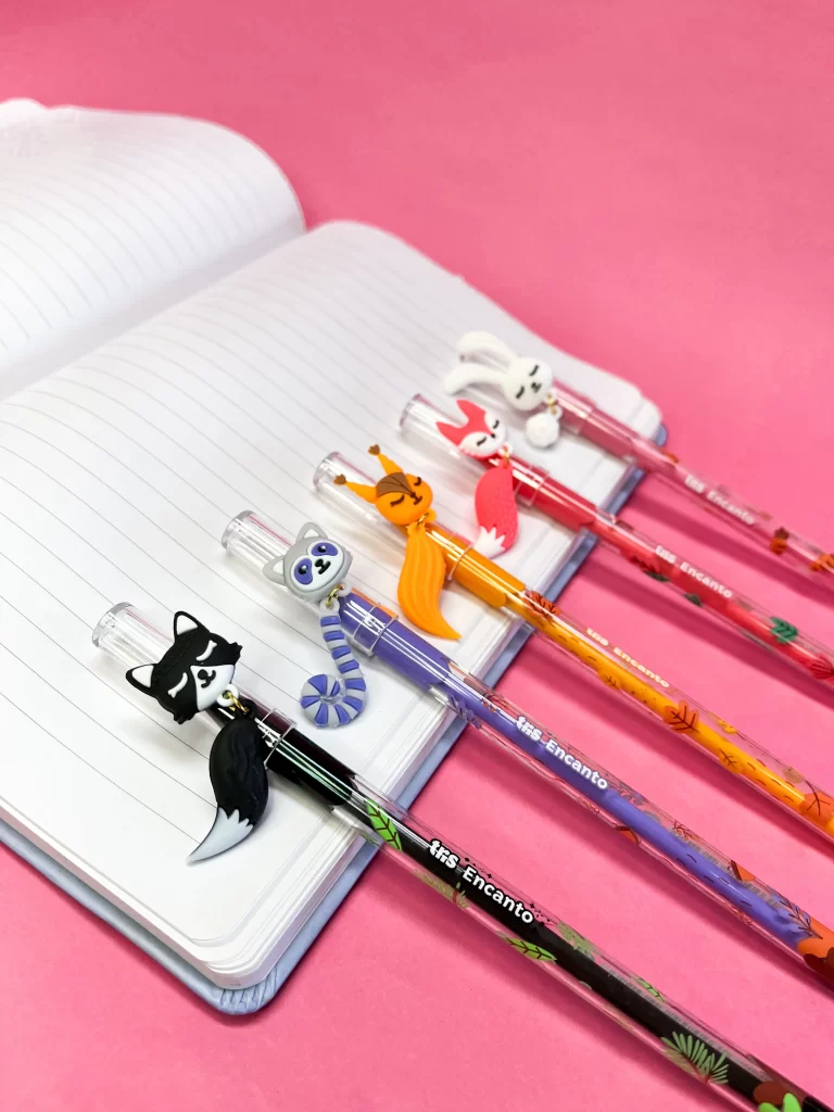 Canetas decoradas com personagens, incluindo uma raposa, um coelho, uma zebra e uma raposa, sobre uma agenda branca com fundo rosa, perfeitas para papelaria fofa e itens escolares.