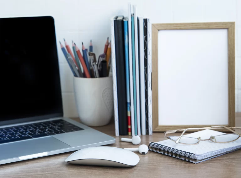 Mesa de escritório com produtos de papelaria, laptop, mouse, livros, quadro branco, óculos e caderno, ambiente de trabalho organizado e ideal para estudos ou trabalho remoto.